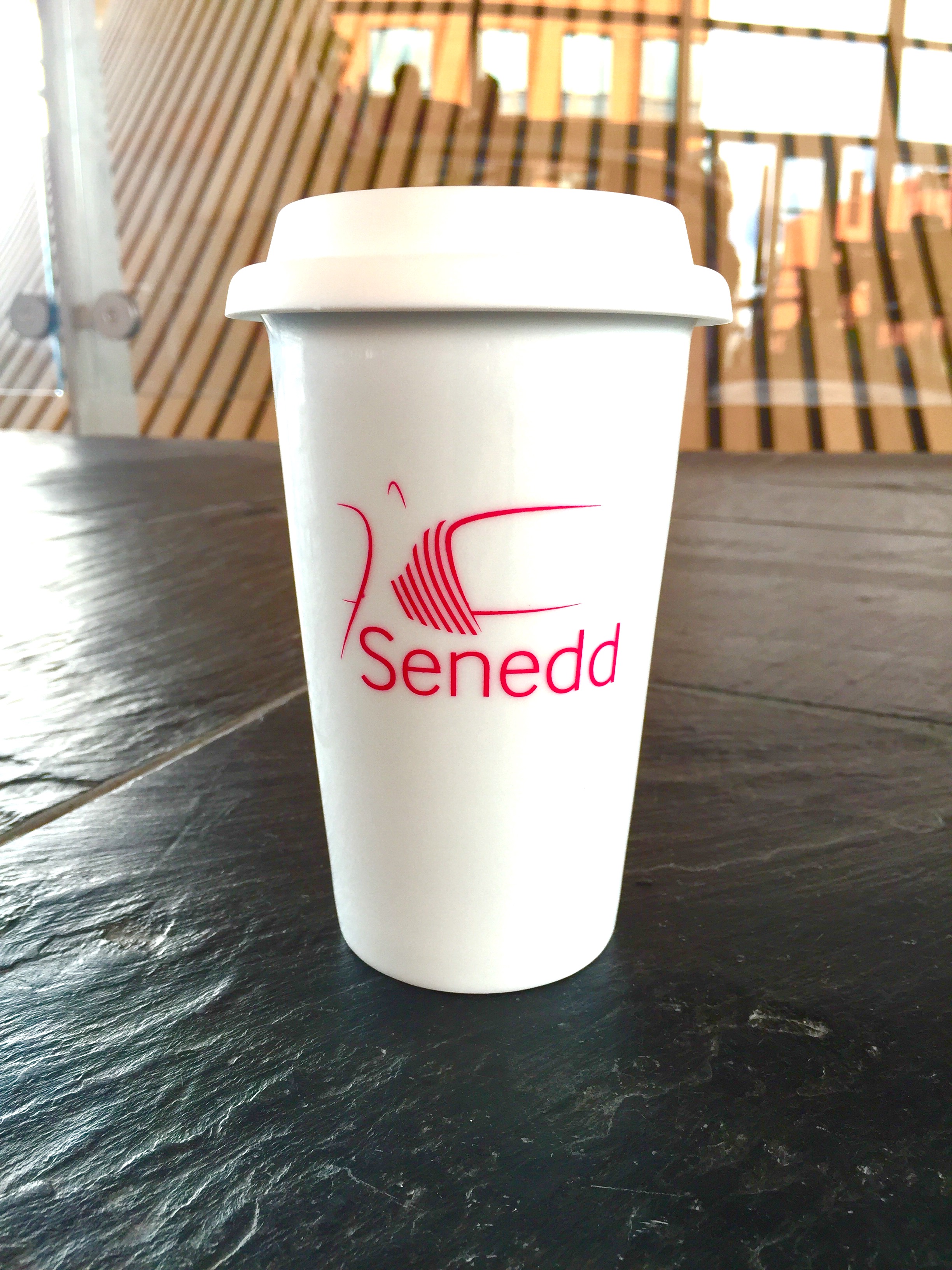 White travel mug with Senedd logo on, the Senedd's funnel in the background.